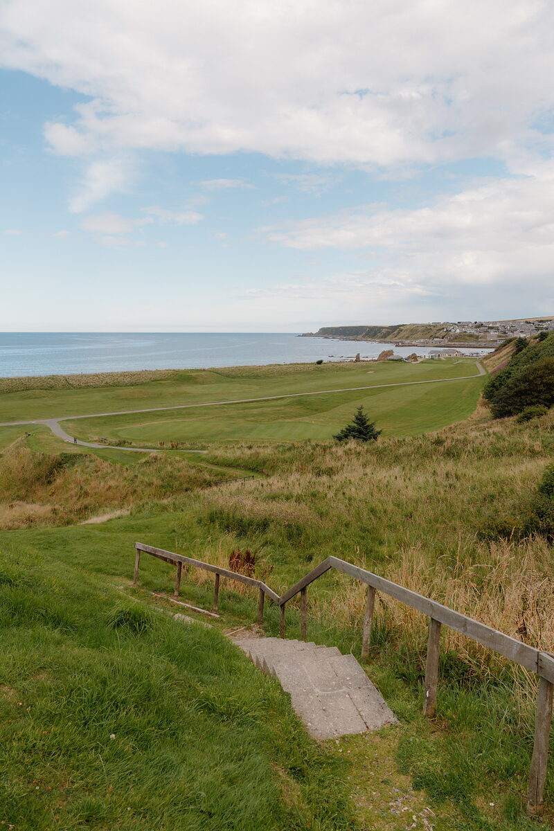 Wooden steps descending through grass towards a fairway and coastline stretching to a distant town
