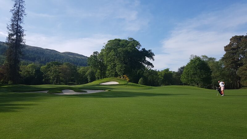 A golfer with a bag on a broad fairway, with deep white bunkers before a raised green, big oaks and a tall conifer, and a wooded hillside behind.