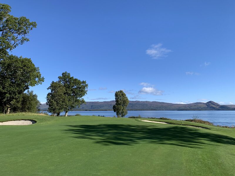 A green with bunkers on either side and Loch Lomond beyond it, with hills along the far shore under a clear blue sky.
