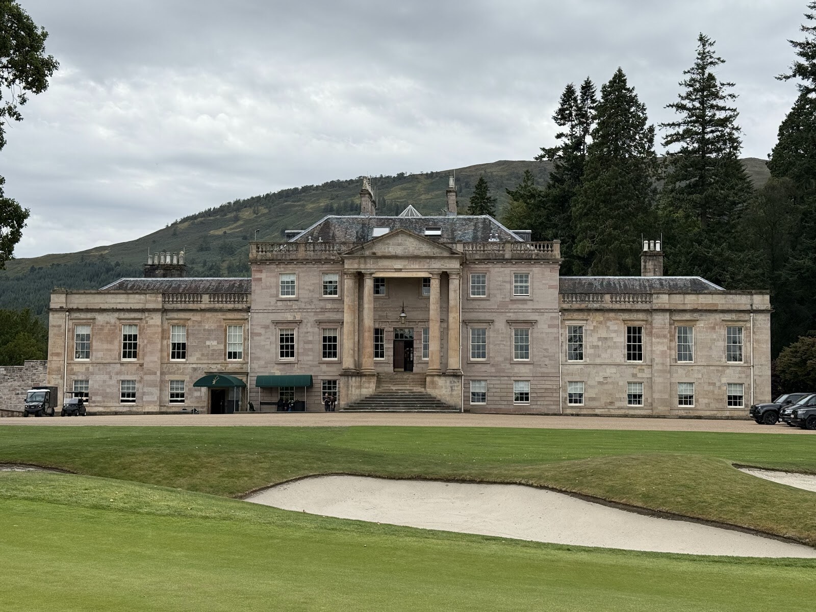 Rossdhu House, the Georgian clubhouse, with its columned portico and steps, seen across a bunker and fairway, with a wooded hill behind.