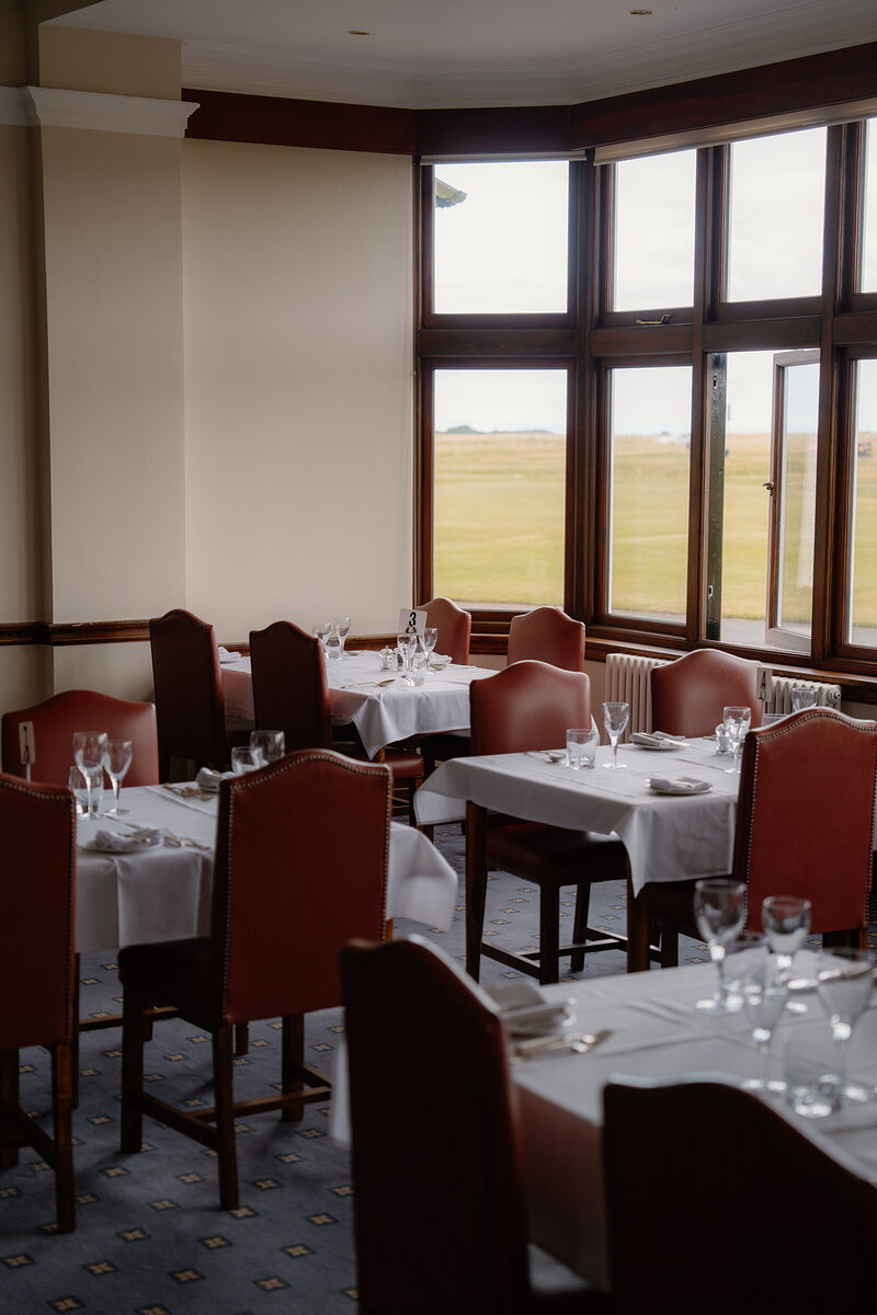 A golf clubhouse dining room with tables set beside bay windows overlooking the links