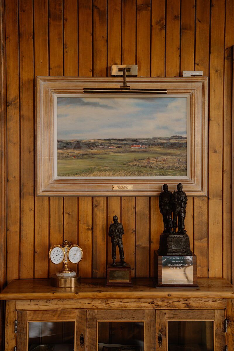 A framed painting of a links course above a sideboard displaying trophies and military figurines