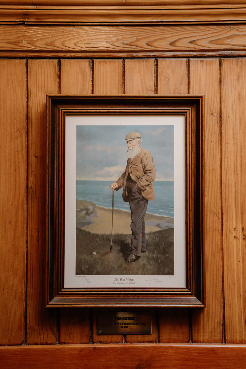 A framed portrait of Old Tom Morris holding a golf club on a beach, mounted on wood panelling with a plaque below