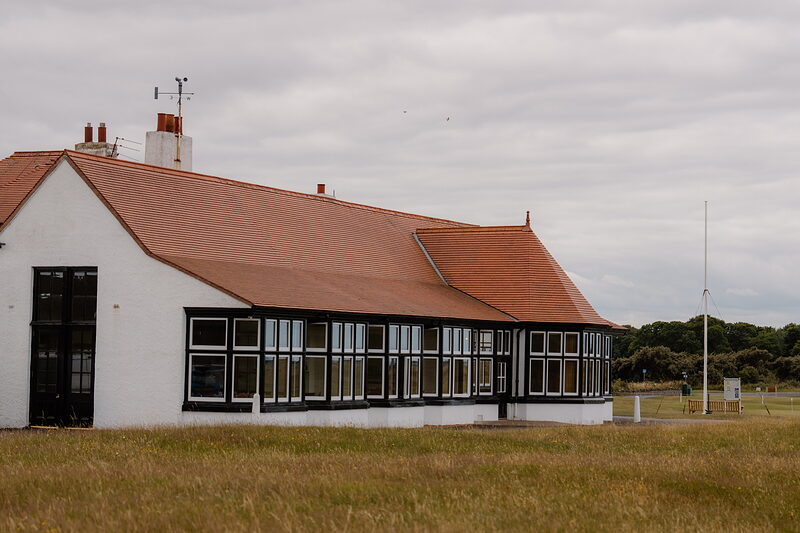A black-and-white timbered clubhouse with a red tiled roof and bay windows beside a flagpole