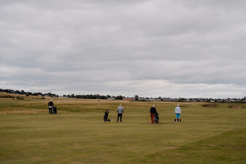 Golfers with trolleys crossing a wide fairway, a village visible on the horizon