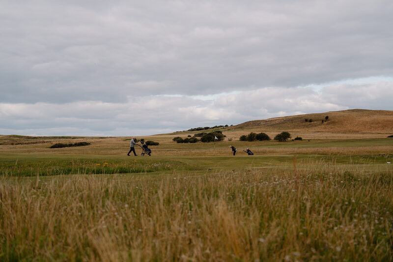 Golfers with trolleys crossing a wide fairway bordered by rolling hills