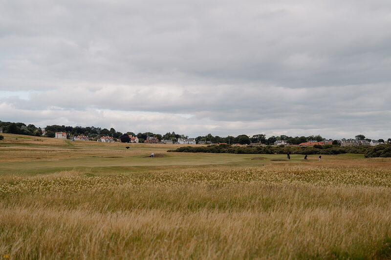 A wide fairway bordered by long grass, a village visible on the horizon with golfers scattered across the course
