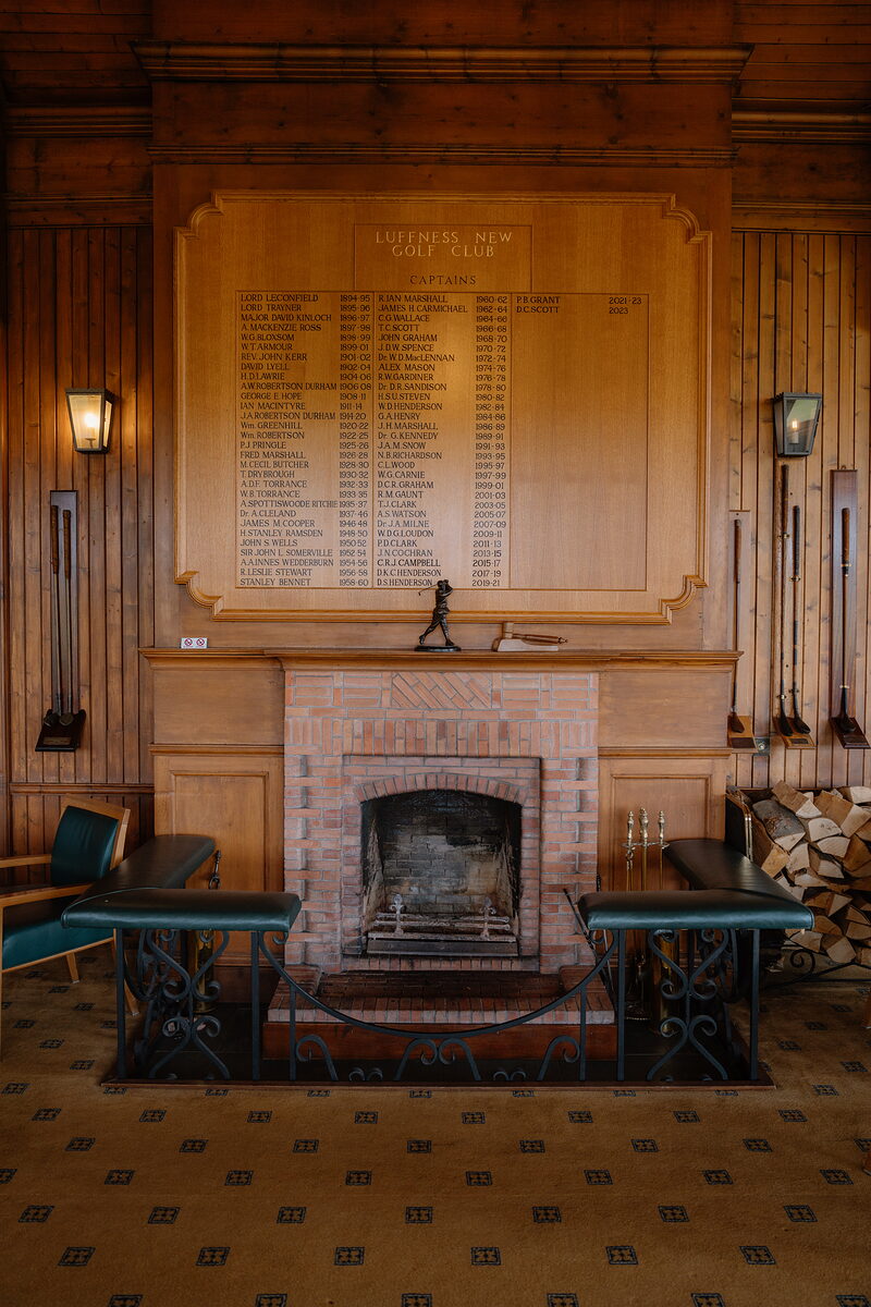 A wood-panelled room with a captains' honours board above a brick fireplace, golf clubs mounted either side