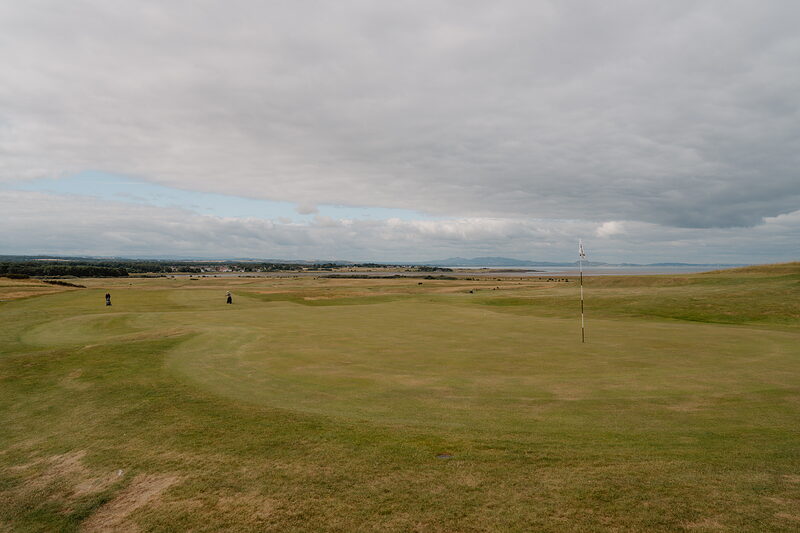 A green with two golfers approaching, a town and firth stretching along the horizon