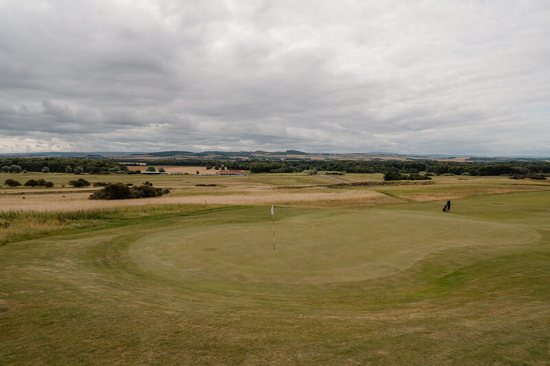 A green marked by a flag before farmland and distant hills, a dark shape standing at its edge