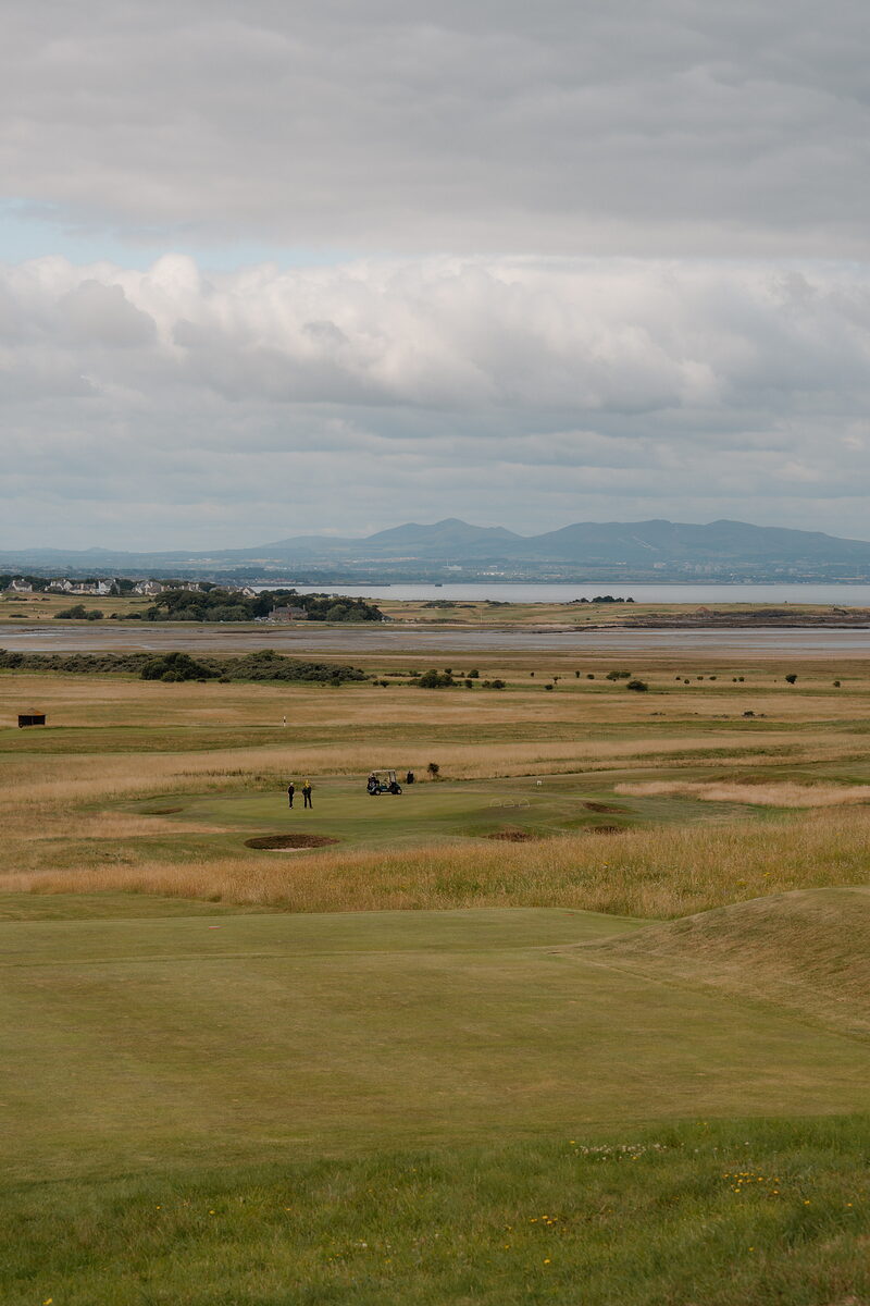 Golfers with a buggy on a green, a firth and distant hills stretching along the horizon