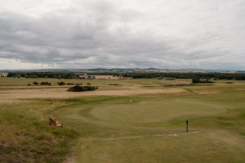 A green marked by a flag before farmland and distant hills, a wooden bench set at its edge
