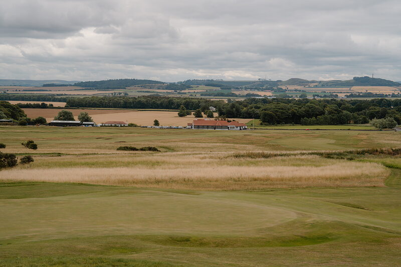 A fairway leading towards a distant clubhouse and farmland stretching to wooded hills