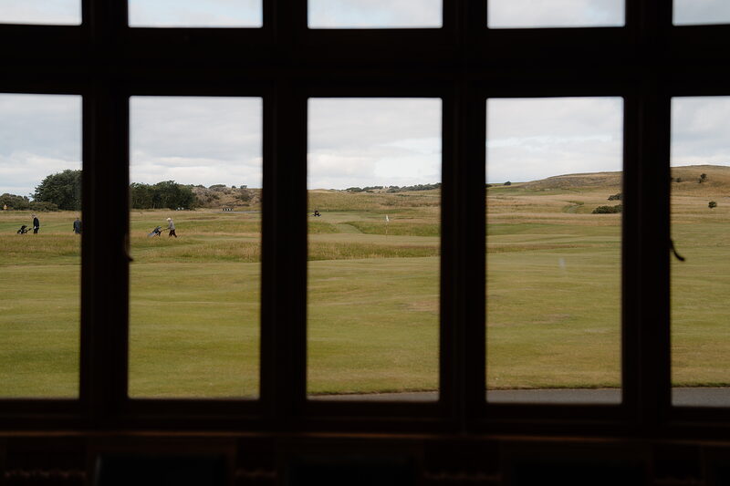 Golfers on a fairway seen through a clubhouse's paned window, hills fading into the distance