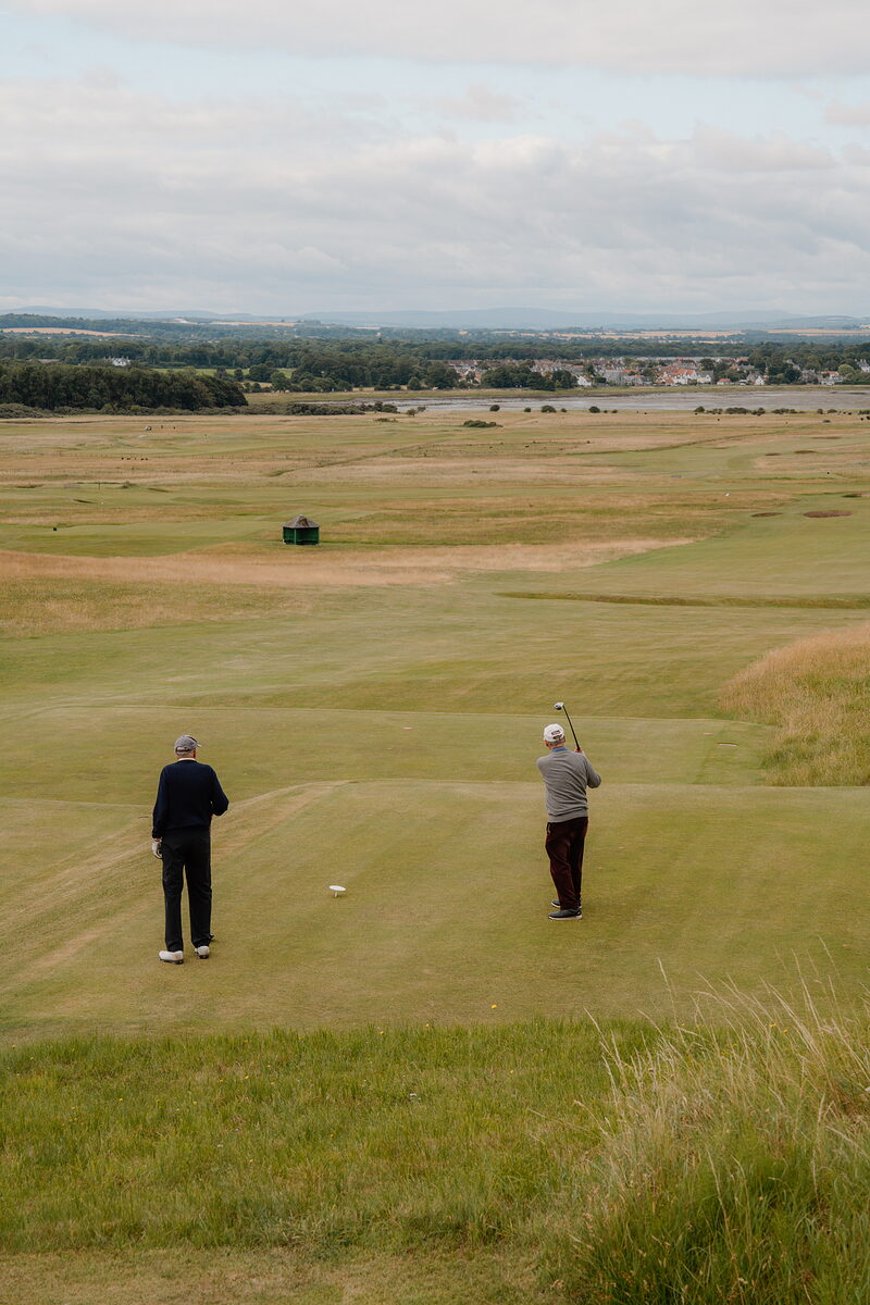 Two golfers on a fairway, one mid-swing, a firth and distant town stretching along the horizon