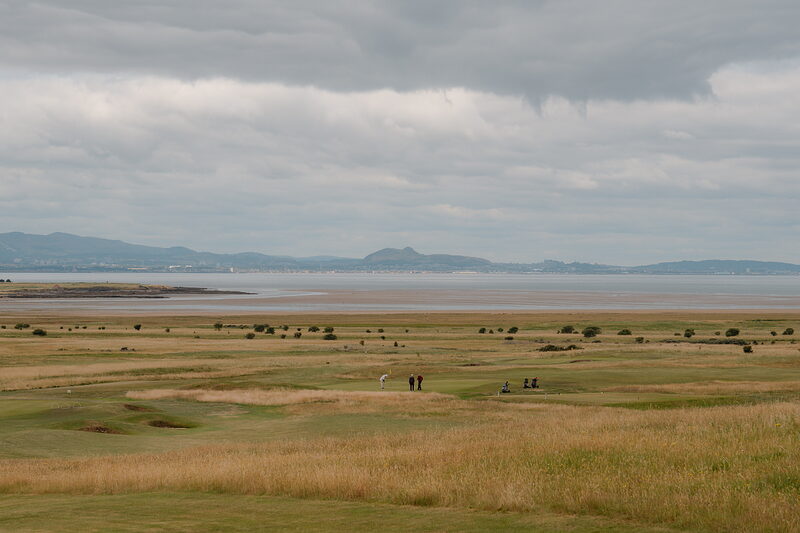 Golfers crossing a marshy fairway, dune grass and hills stretching to the horizon