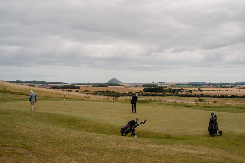 Two golfers with bags standing on a green, a conical hill rising above farmland in the distance
