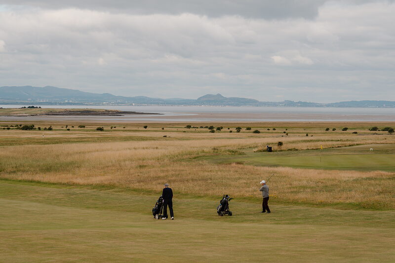 Golfers with bags standing on a fairway, marshland and hills stretching to the horizon