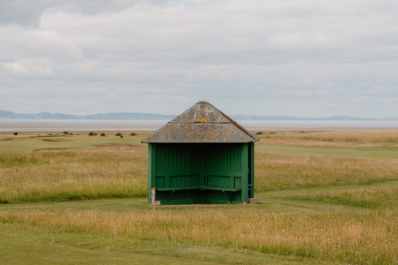 A green timber shelter with a pitched roof standing alone on a fairway, a firth stretching into the distance
