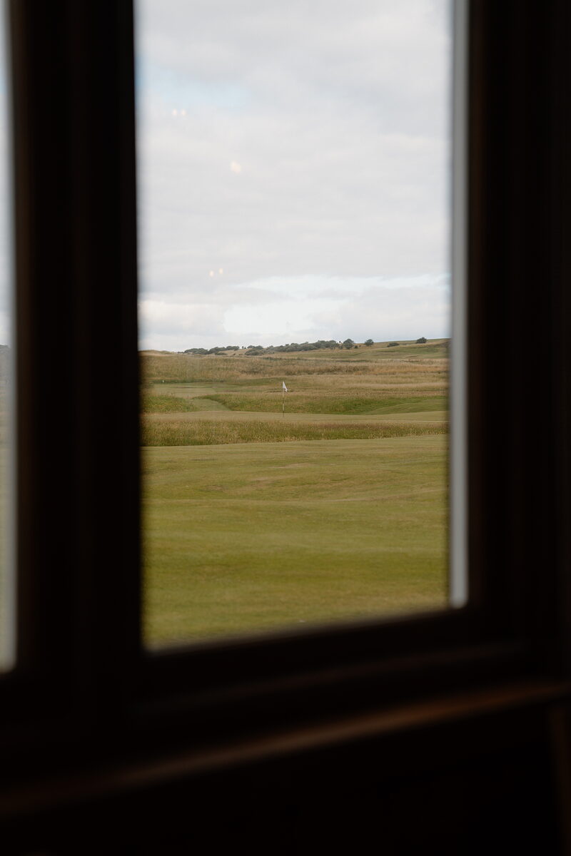 A green marked by a white flag seen through a paned window, rough grass framing the fairway