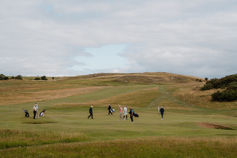 Golfers gathered on a fairway with bags and trolleys, a hill rising beyond the gorse