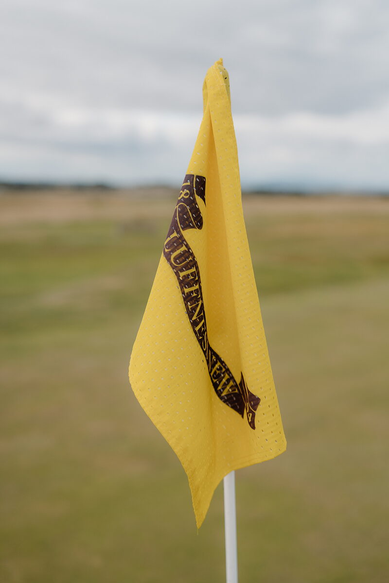 A yellow flag bearing the Luffness New crest fluttering above a fairway of rough grass