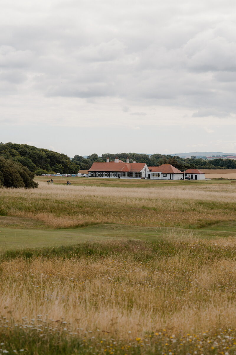 A golfer with a bag walking a fairway before a red-roofed clubhouse