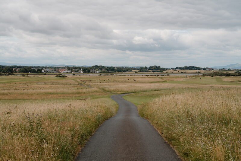 A tarmac path curving through farmland towards a distant village under a heavy sky.