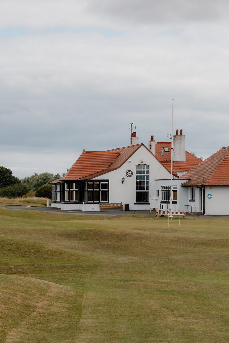 A white clubhouse with a clock and orange-tiled roof, flags marking greens on the fairway before it