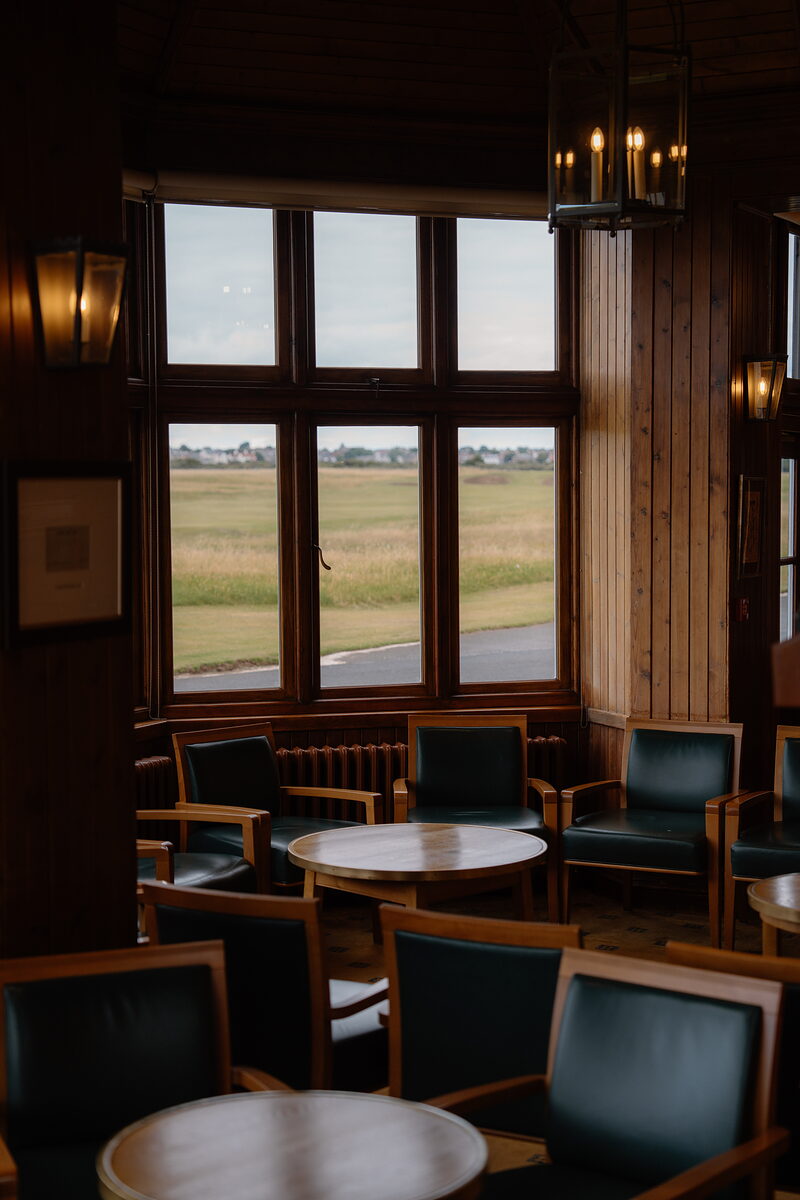 A clubhouse lounge with green leather armchairs and round tables set beneath tall windows overlooking the course