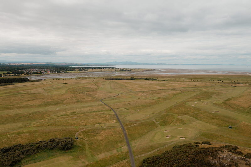 A fairway winding towards a firth and distant hills, a road cutting through the mown grass