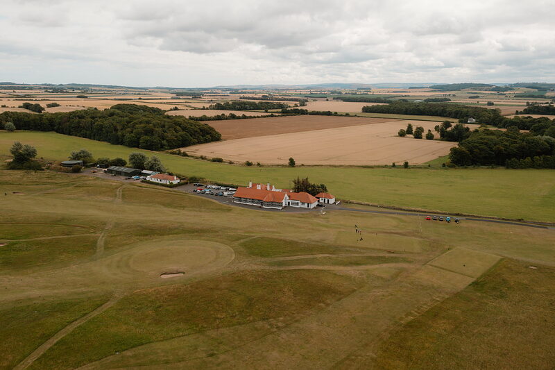 A row of sandstone clubhouse buildings set before farmland stretching to the horizon