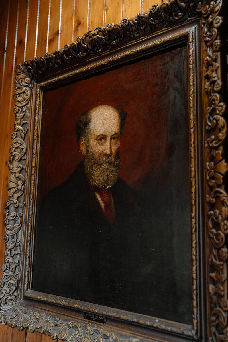 A framed oil portrait of a bearded man in a dark coat and red cravat, mounted on wood panelling