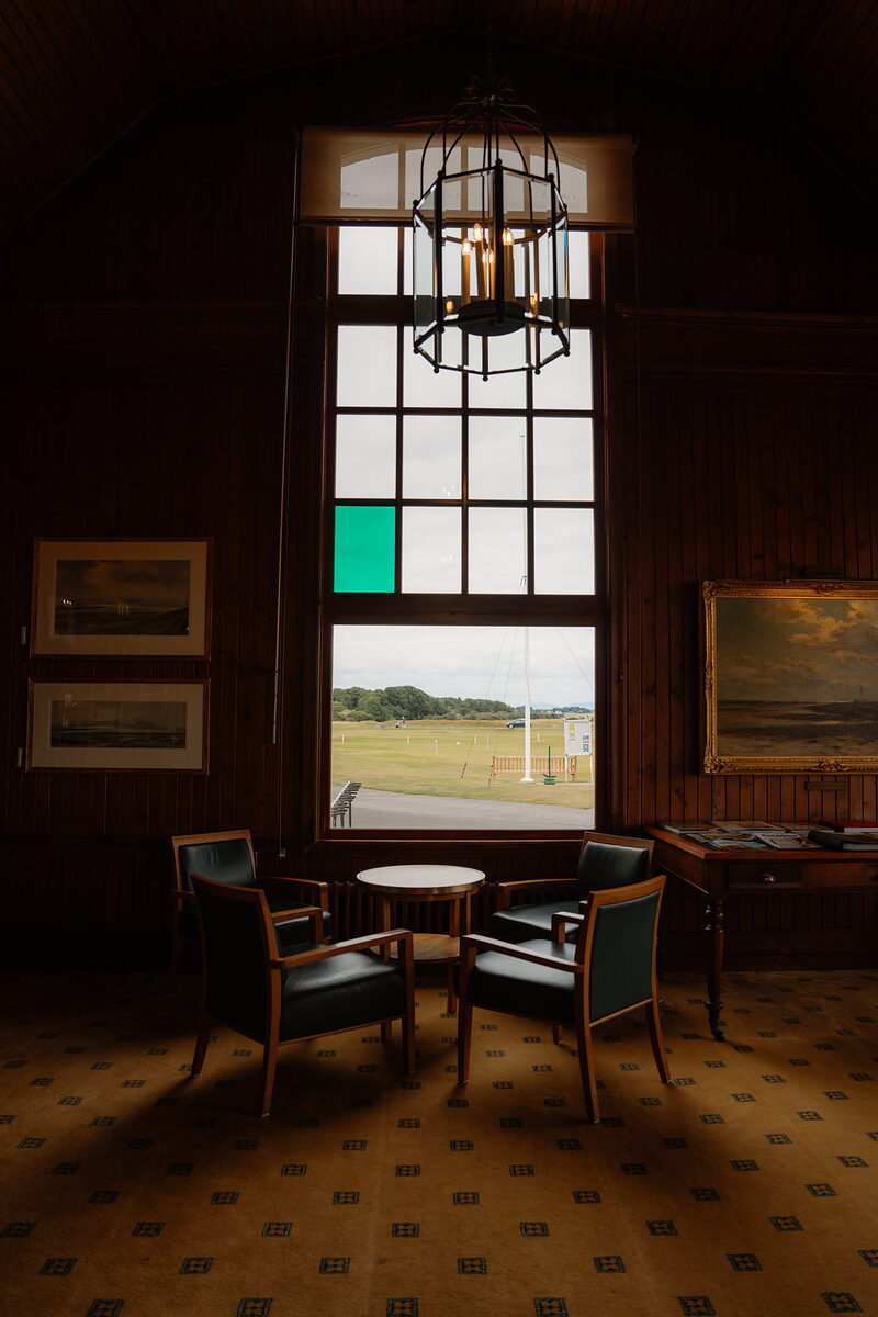A tall paned window with a green glass insert, armchairs and a round table set beneath a lantern chandelier
