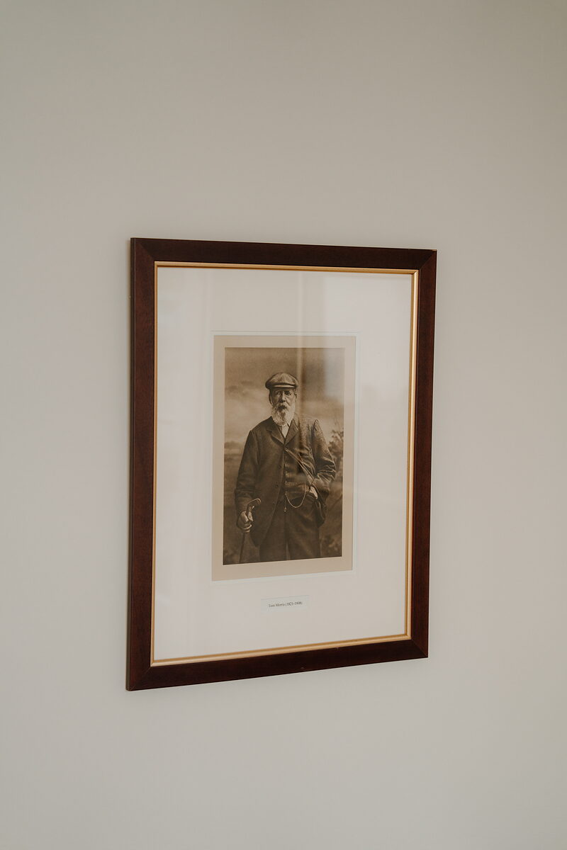 A framed sepia photograph of a bearded golfer in a flat cap holding a club, hung on a plain wall