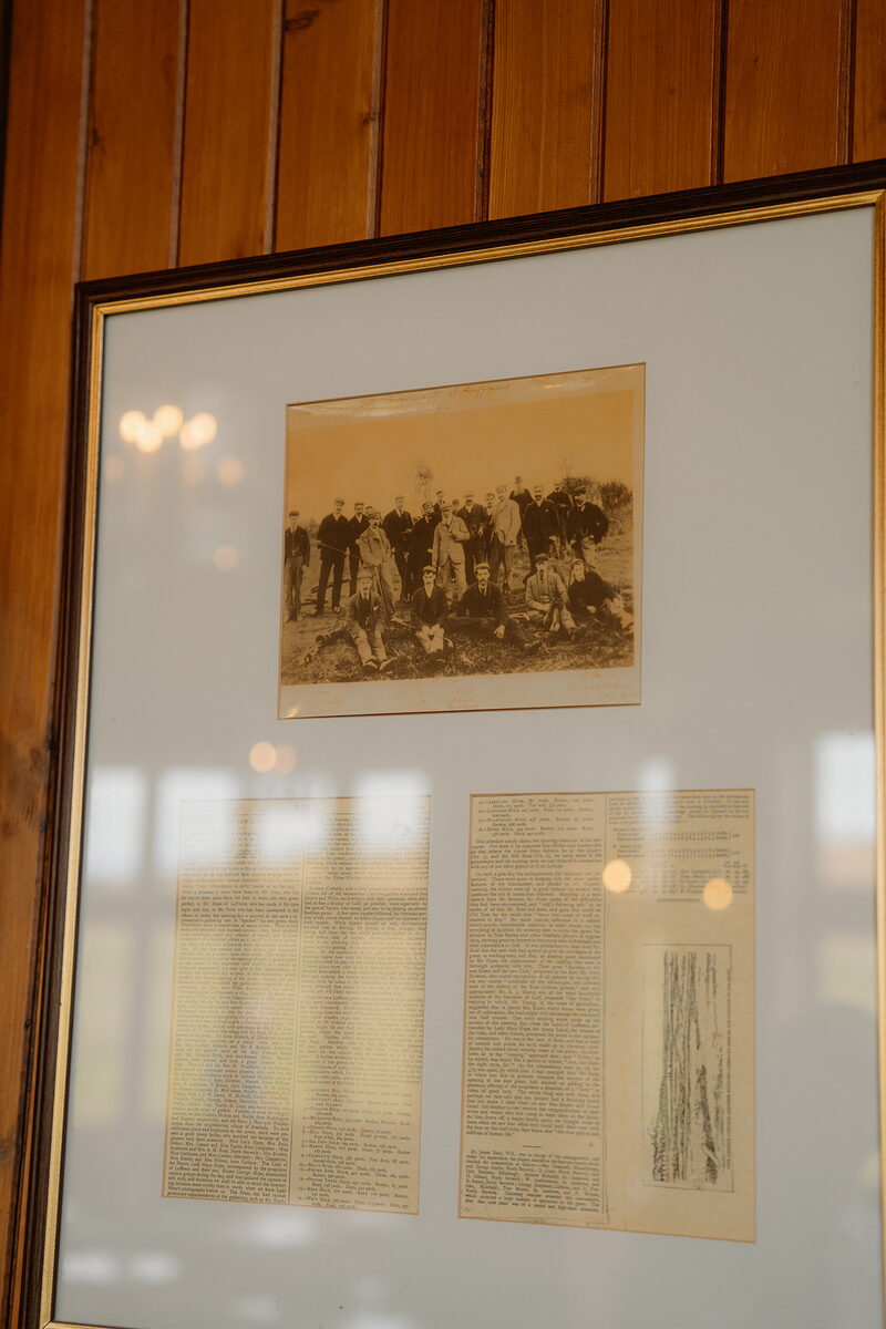 A framed sepia photograph of a golfing party with newspaper clippings mounted beneath it on wood panelling