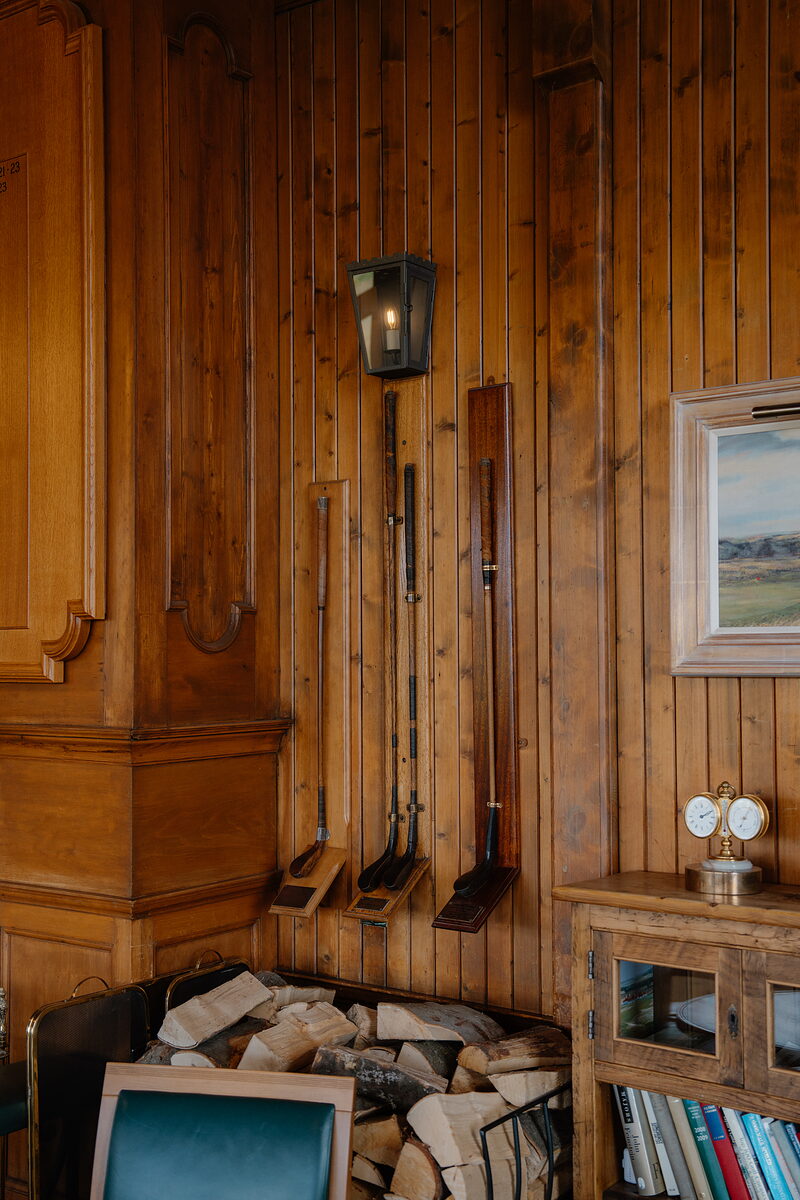 Golf clubs and a lantern mounted on wood-panelled walls beside a framed landscape painting and stacked firewood