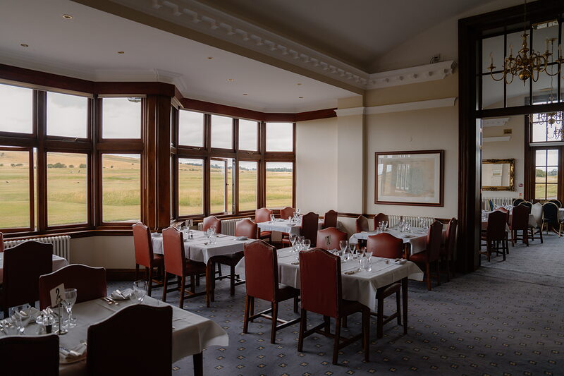 A dining room with red upholstered chairs and tables set beneath bay windows overlooking the course