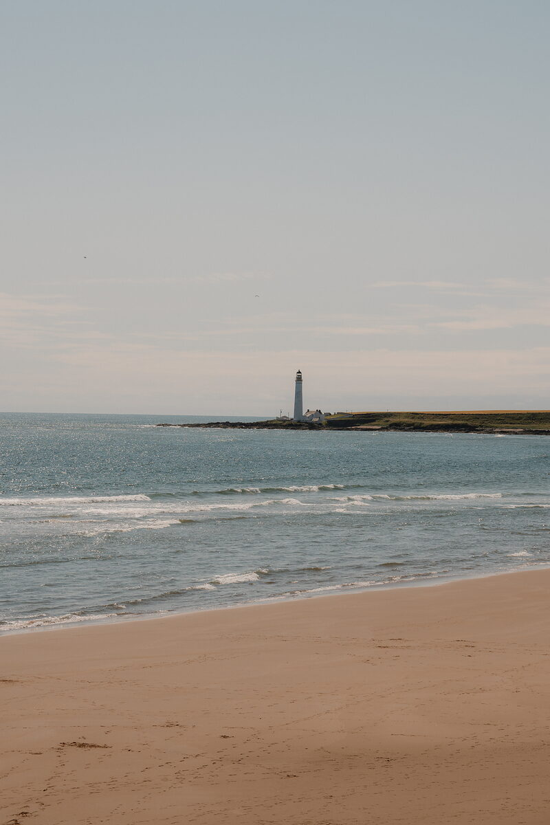 A lighthouse standing on a headland beyond a sandy beach, waves breaking along the shore.
