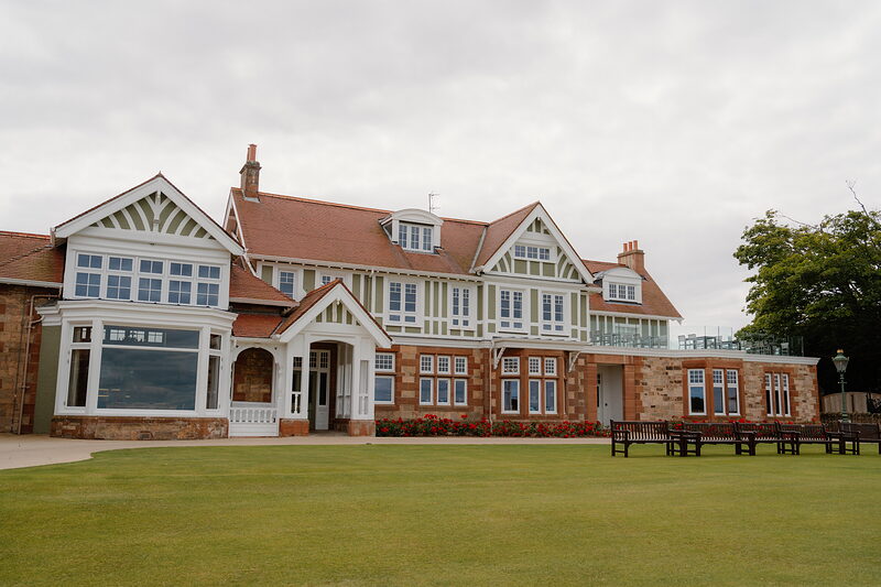 A red sandstone clubhouse with white gables and bay windows overlooking a lawn and benches