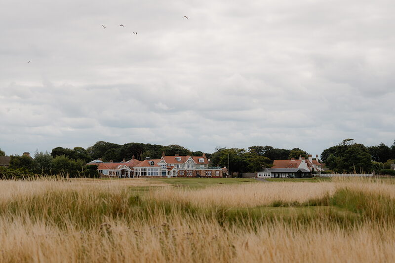 A distant view of a clubhouse and outbuildings across a golf course, birds flying overhead