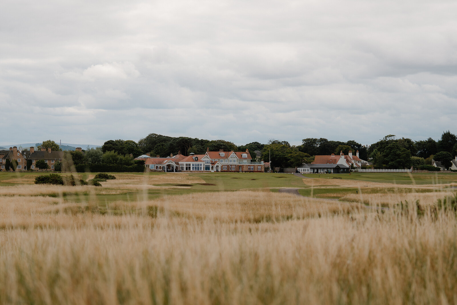 A distant clubhouse and outbuildings seen across golden dune grass on a links course