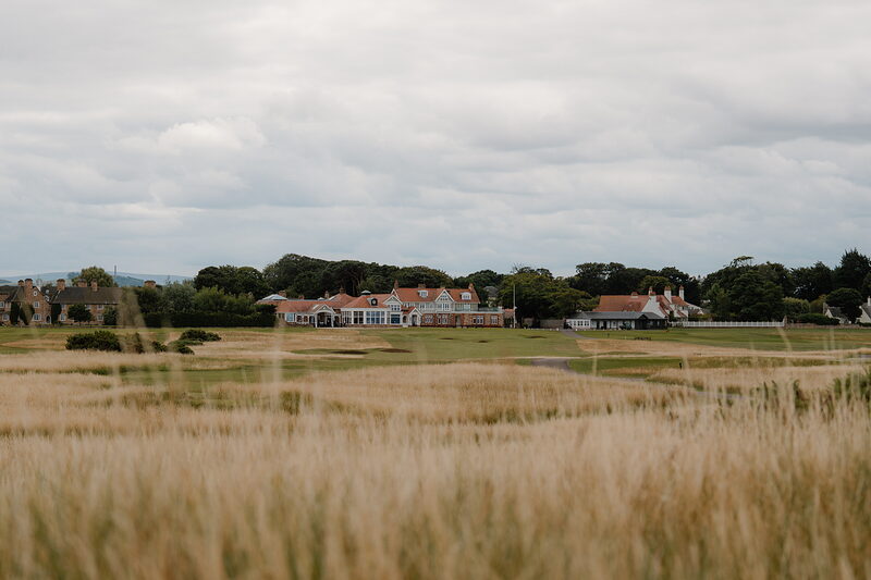 A distant clubhouse and outbuildings seen across golden dune grass on a links course