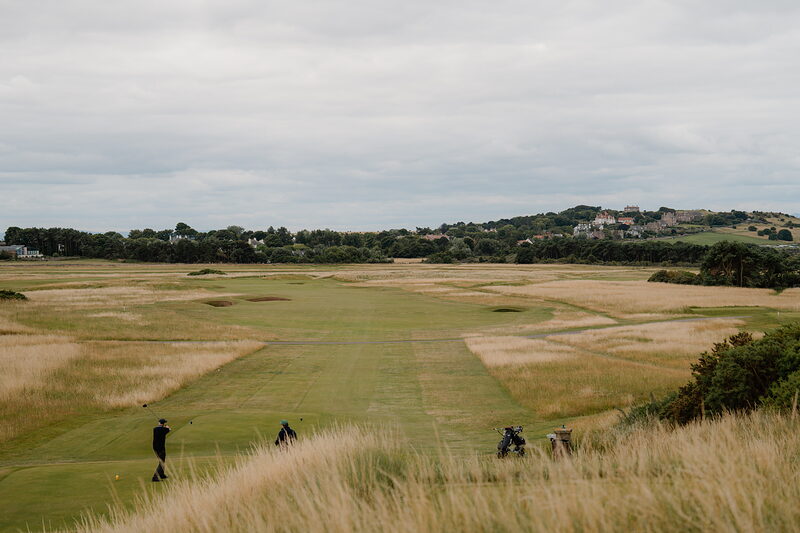 Golfers with trolleys crossing a wide fairway, a village and clubhouse visible on the horizon