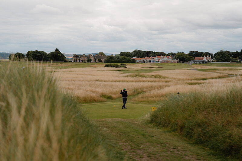 A golfer photographing the view from a tee, a clubhouse and cottages visible across the fairway