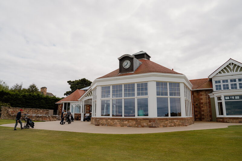 A clubhouse with a clock tower and bay window, golfers gathered with trolleys on the forecourt