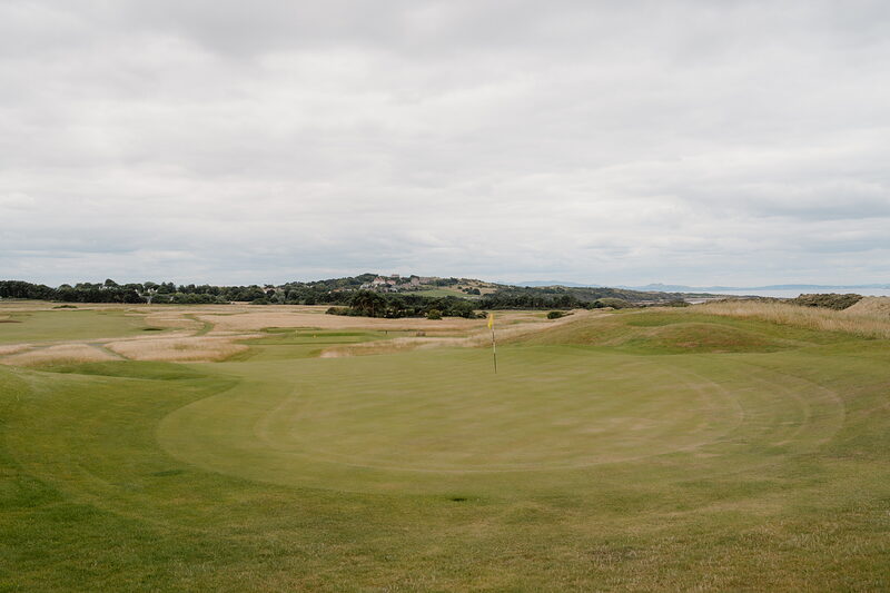 A fairway leading to a green with a clubhouse and hills rising beyond
