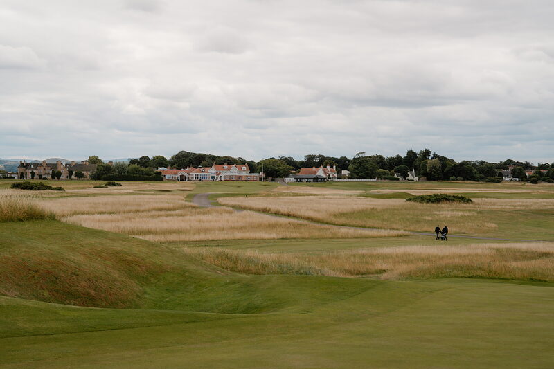 Golfers walking a long grass-lined fairway, a village visible on the horizon