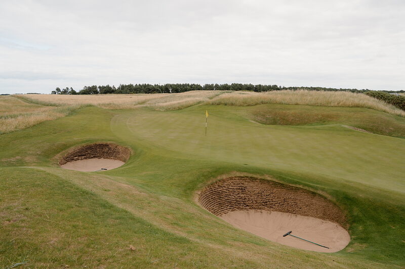 Deep bunkers flanking a green bordered by dune grass and a line of trees
