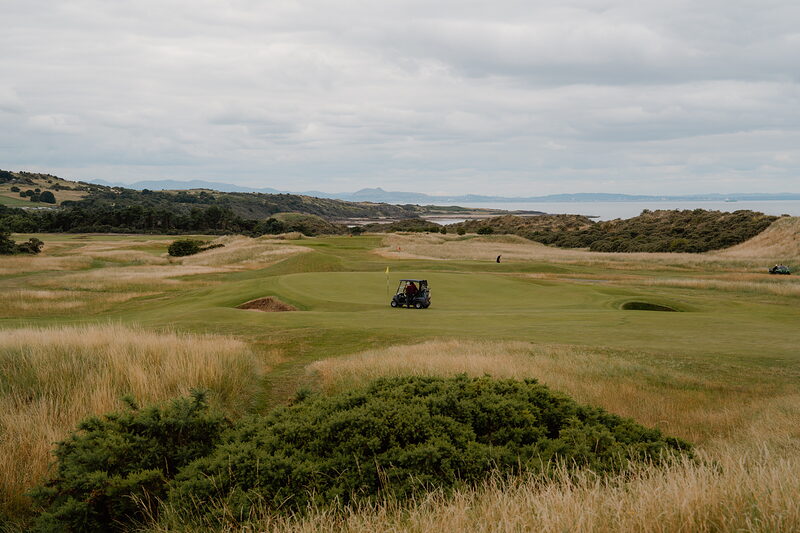 A golf buggy parked on a fairway overlooking dune grass and a bay in the distance
